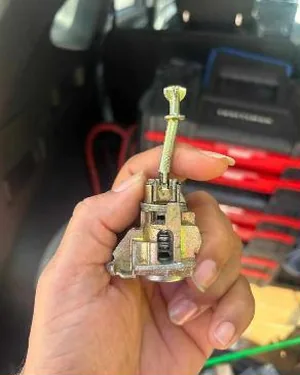 A hand holding a lock cylinder taken apart, with the wafers visible, beside a vehicle and tools.
