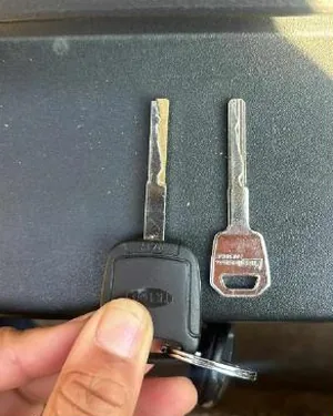 A hand holding two car keys — a black transponder fob and a freshly cut silver key — over a dark car interior.
