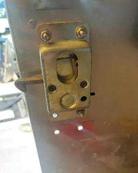 Close-up of a brass latch mechanism on the edge of an aluminium door, photographed outdoors in daylight.