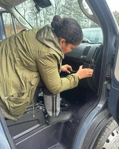 A Dee Locksmith locksmith in an olive jacket leaning into the open driver's door of an SUV, working on the door panel.